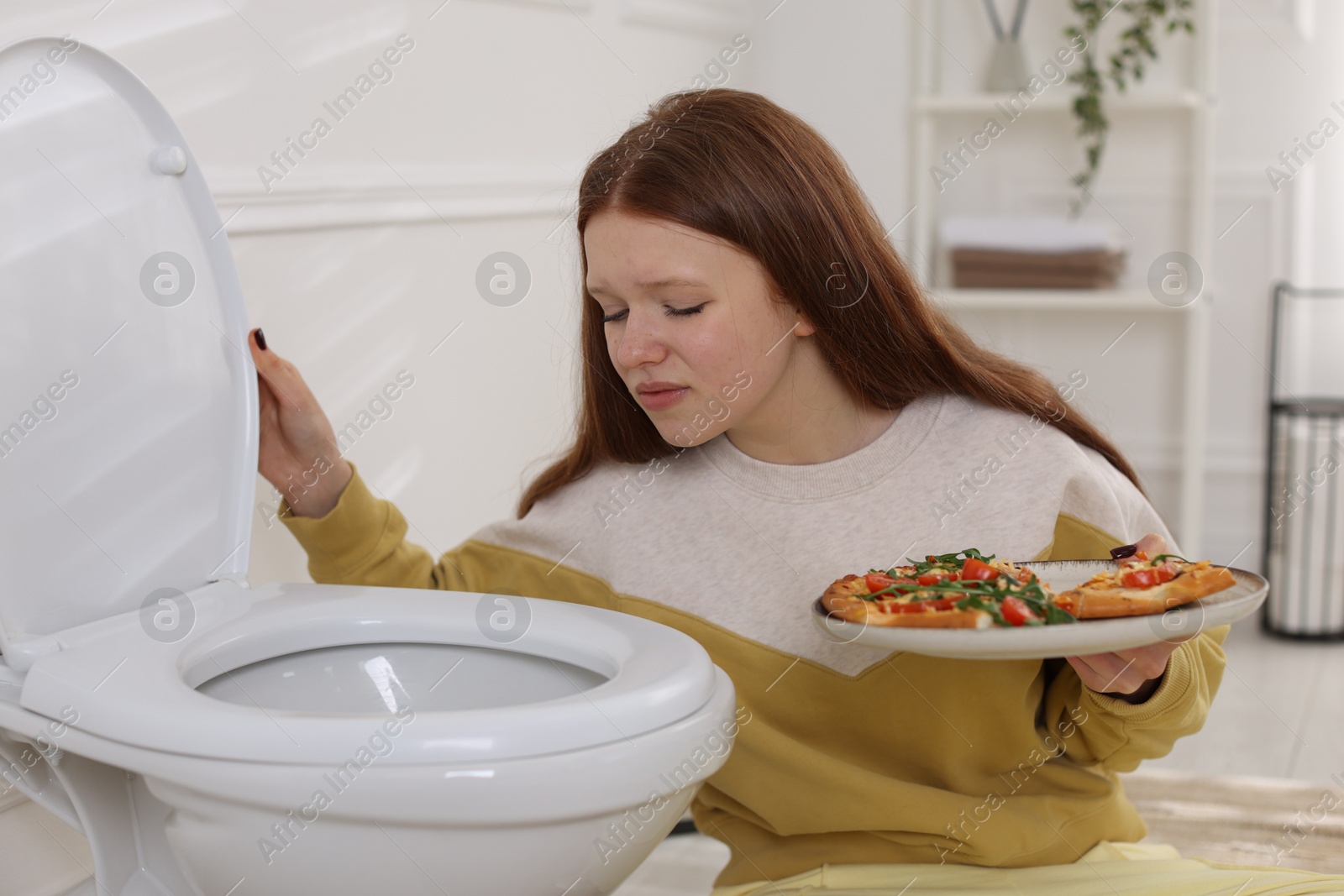 Photo of Sad teenage girl with pizza near toilet bowl in bathroom. Bulimia