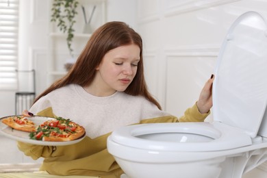 Photo of Sad teenage girl with pizza near toilet bowl in bathroom. Bulimia