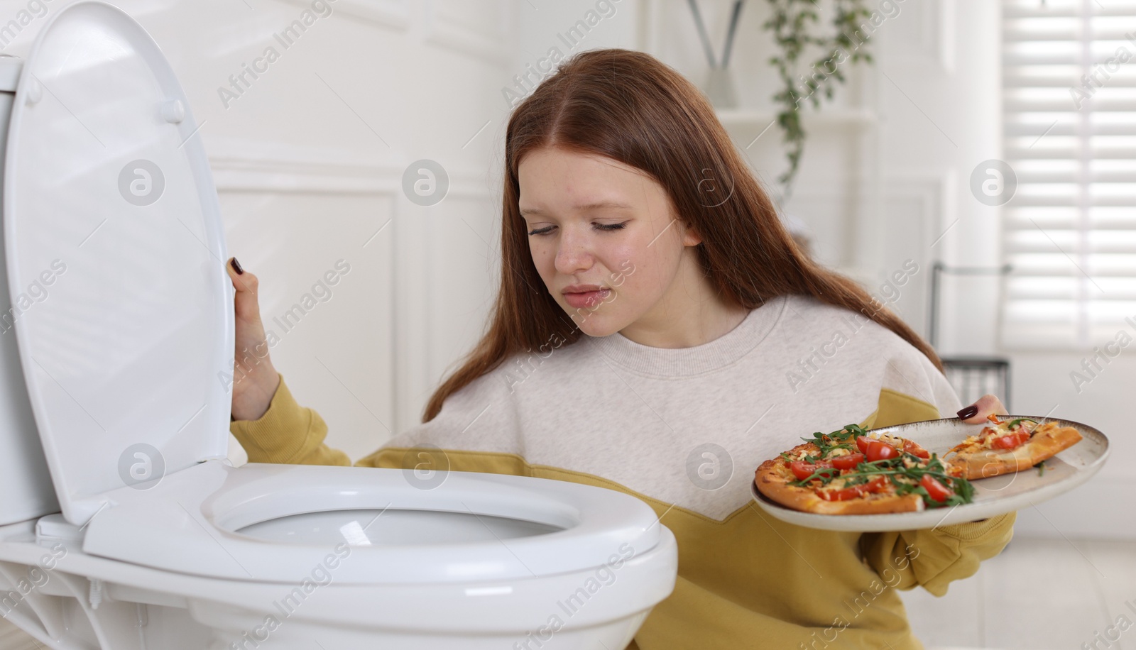 Photo of Sad teenage girl with pizza near toilet bowl in bathroom. Bulimia