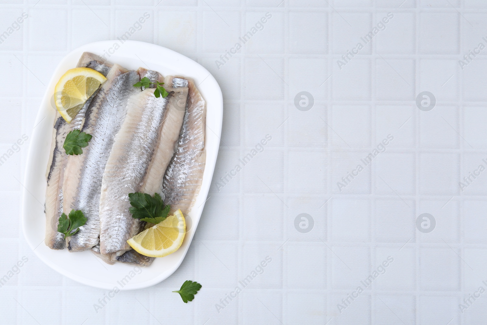 Photo of Marinated herring fillets with lemon and parsley on white tiled table, top view. Space for text