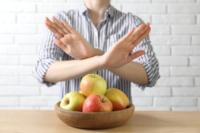 Photo of Woman refusing to eat apples at wooden table near white brick wall, selective focus. Food allergy concept