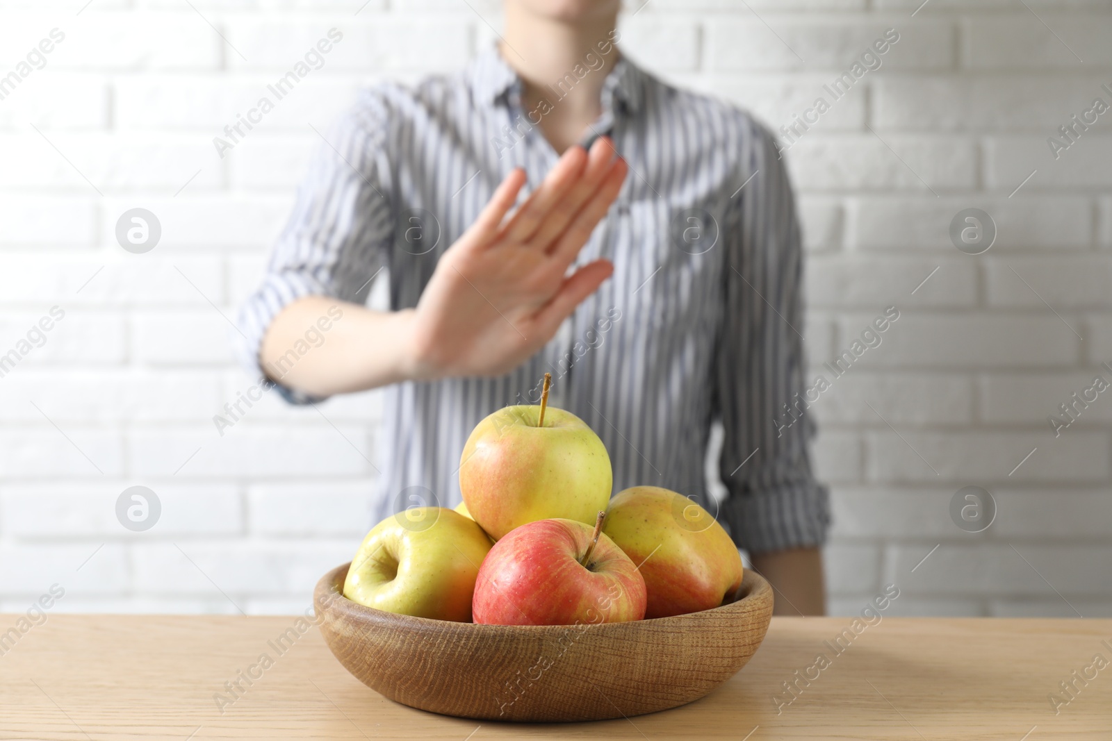 Photo of Woman refusing to eat apples at wooden table near white brick wall, selective focus. Food allergy concept