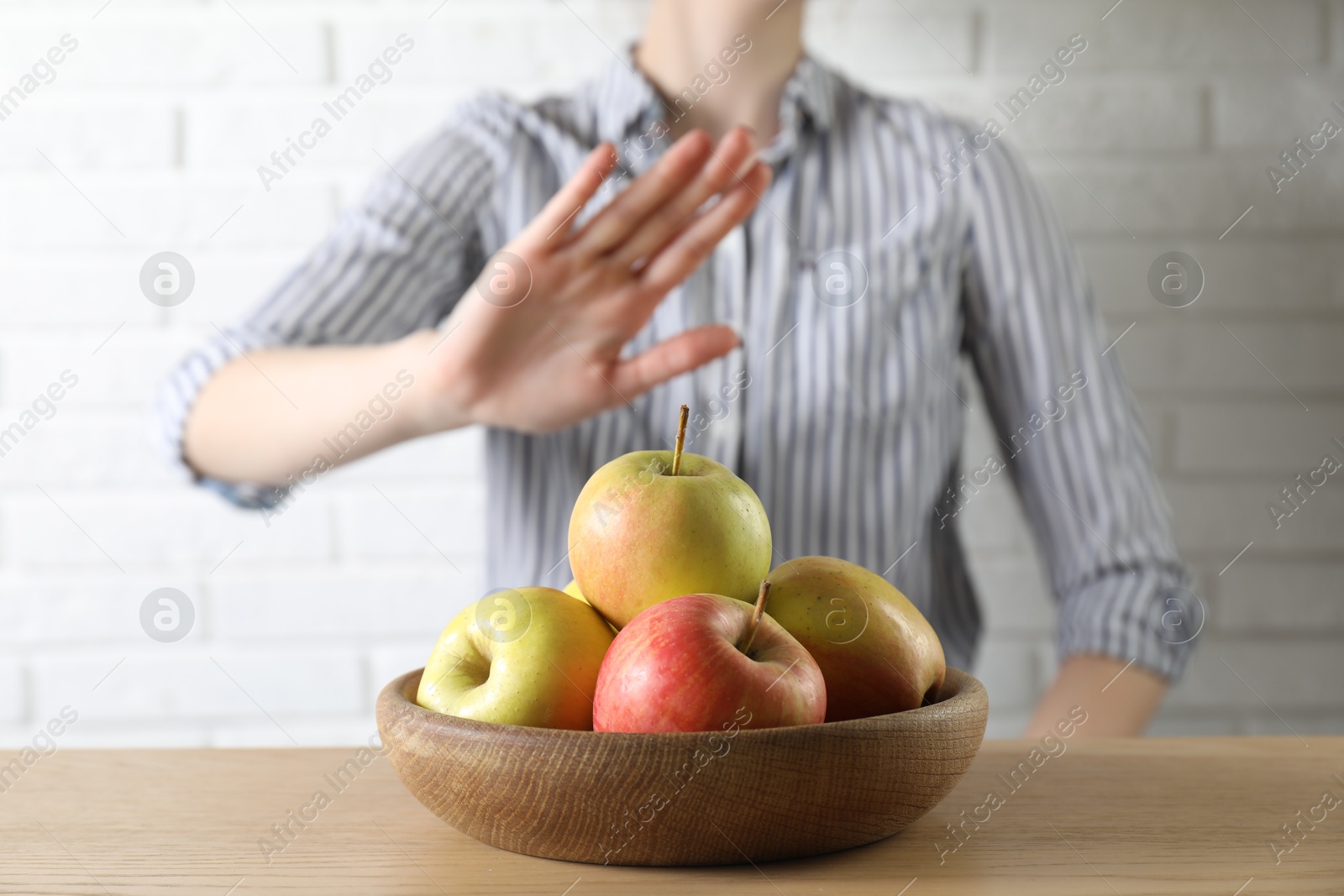 Photo of Woman refusing to eat apples at wooden table near white brick wall, selective focus. Food allergy concept