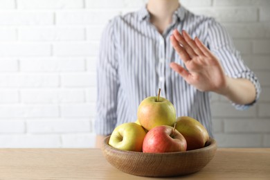 Photo of Woman refusing to eat apples at wooden table near white brick wall, selective focus. Food allergy concept