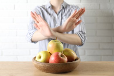 Photo of Woman refusing to eat apples at wooden table near white brick wall, selective focus. Food allergy concept