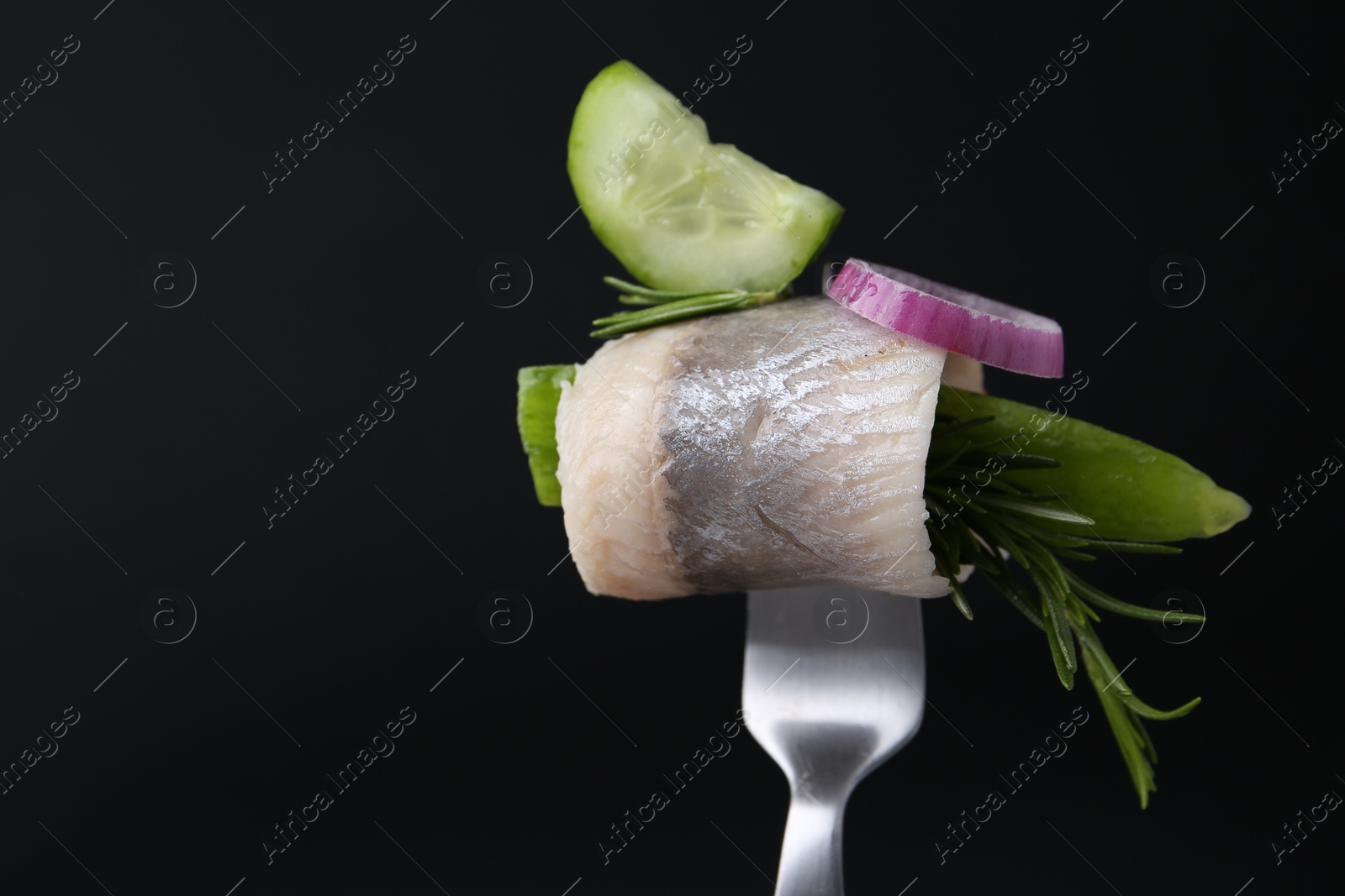 Photo of Fork with marinated herring fillet, cucumber, rosemary and onion ring on black background, closeup. Space for text