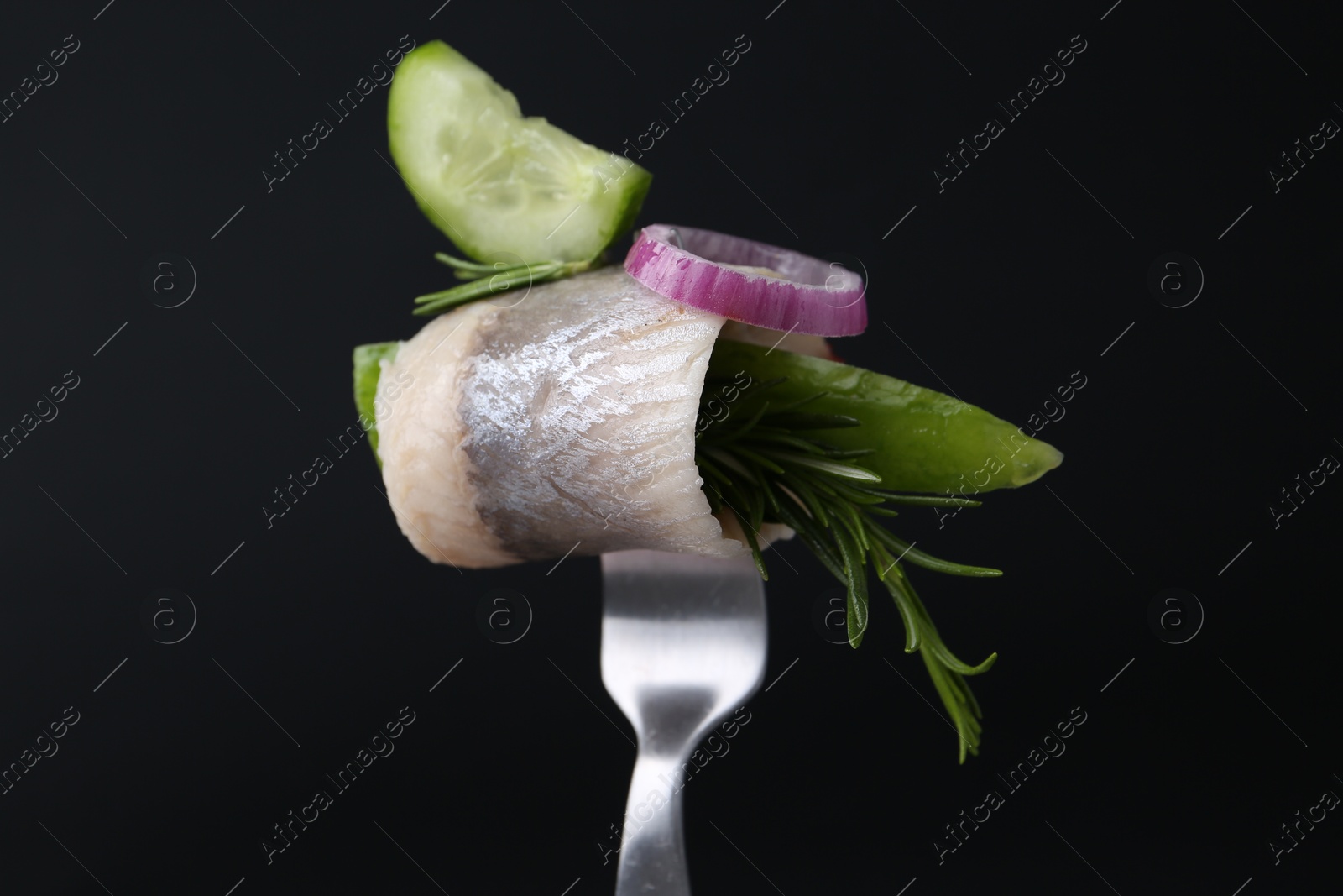 Photo of Fork with marinated herring fillet, cucumber, rosemary and onion ring on black background, closeup