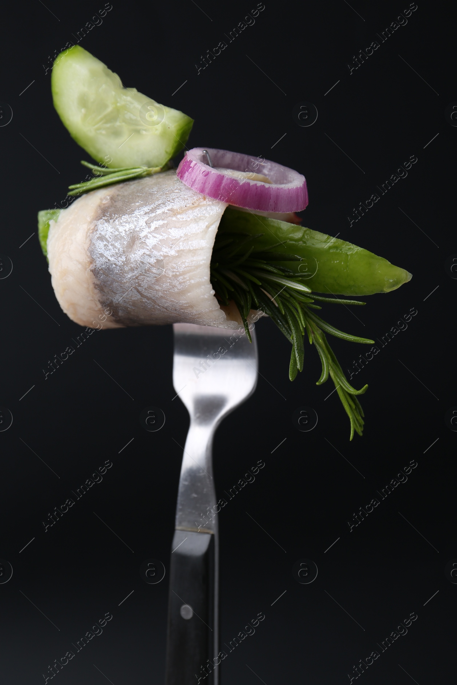 Photo of Fork with marinated herring fillet, cucumber, rosemary and onion ring on black background, closeup