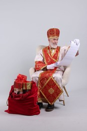 Photo of Saint Nicholas with wish list and sack full of gifts on light grey background