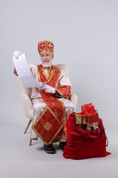 Photo of Saint Nicholas with wish list and sack full of gifts on light grey background