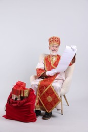 Saint Nicholas with wish list and sack full of gifts on light grey background