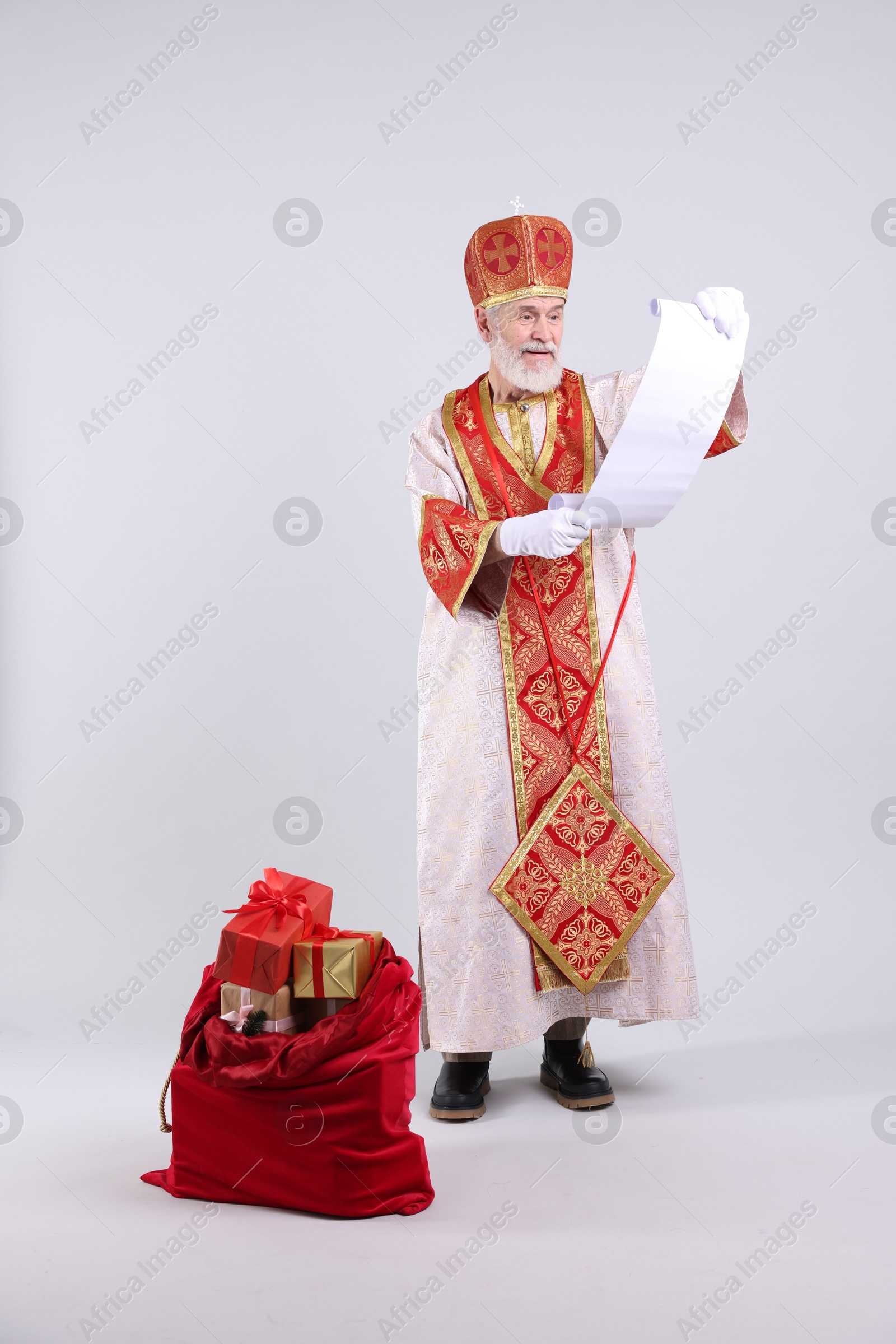 Photo of Saint Nicholas with wish list and sack full of gifts on light grey background