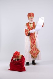 Photo of Saint Nicholas with wish list and sack full of gifts on light grey background