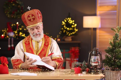 Photo of Saint Nicholas with notebook at desk in room decorated for Christmas