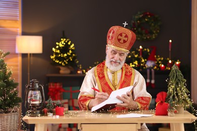 Photo of Saint Nicholas with notebook at desk in room decorated for Christmas