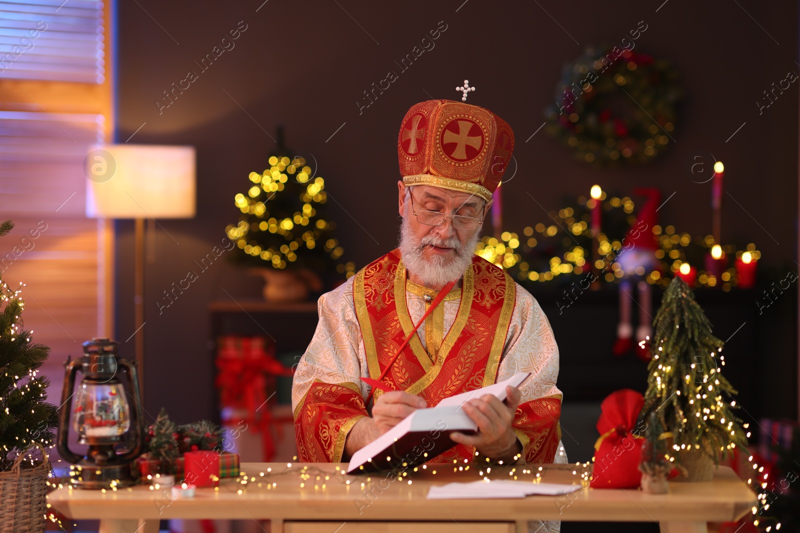 Photo of Saint Nicholas with notebook at desk in room decorated for Christmas