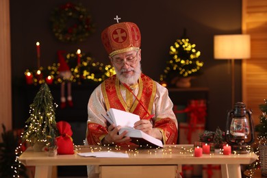 Saint Nicholas with notebook at desk in room decorated for Christmas