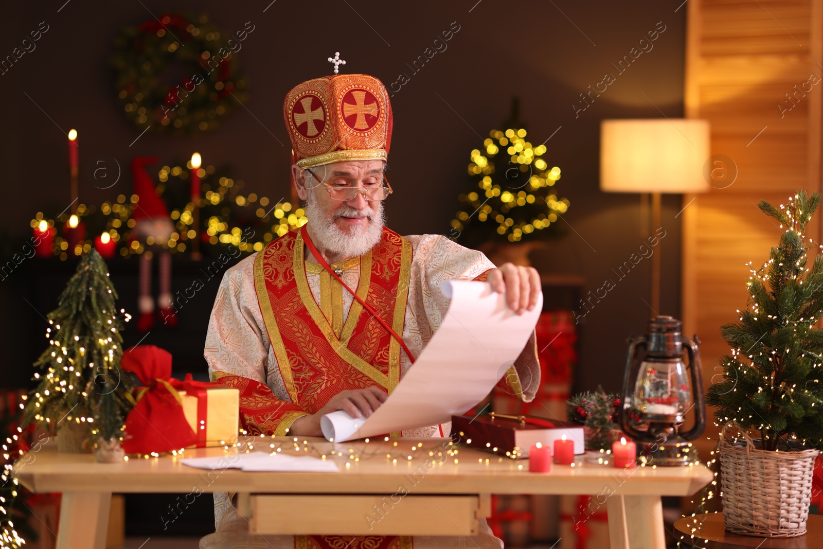 Photo of Saint Nicholas with gift list at desk in room decorated for Christmas