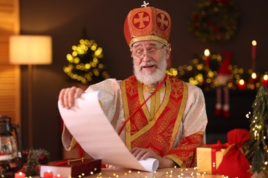 Photo of Saint Nicholas with gift list at desk in room decorated for Christmas