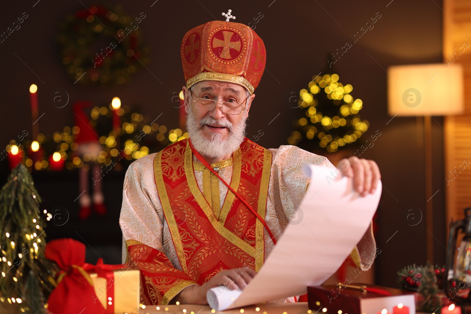 Photo of Saint Nicholas with gift list at desk in room decorated for Christmas