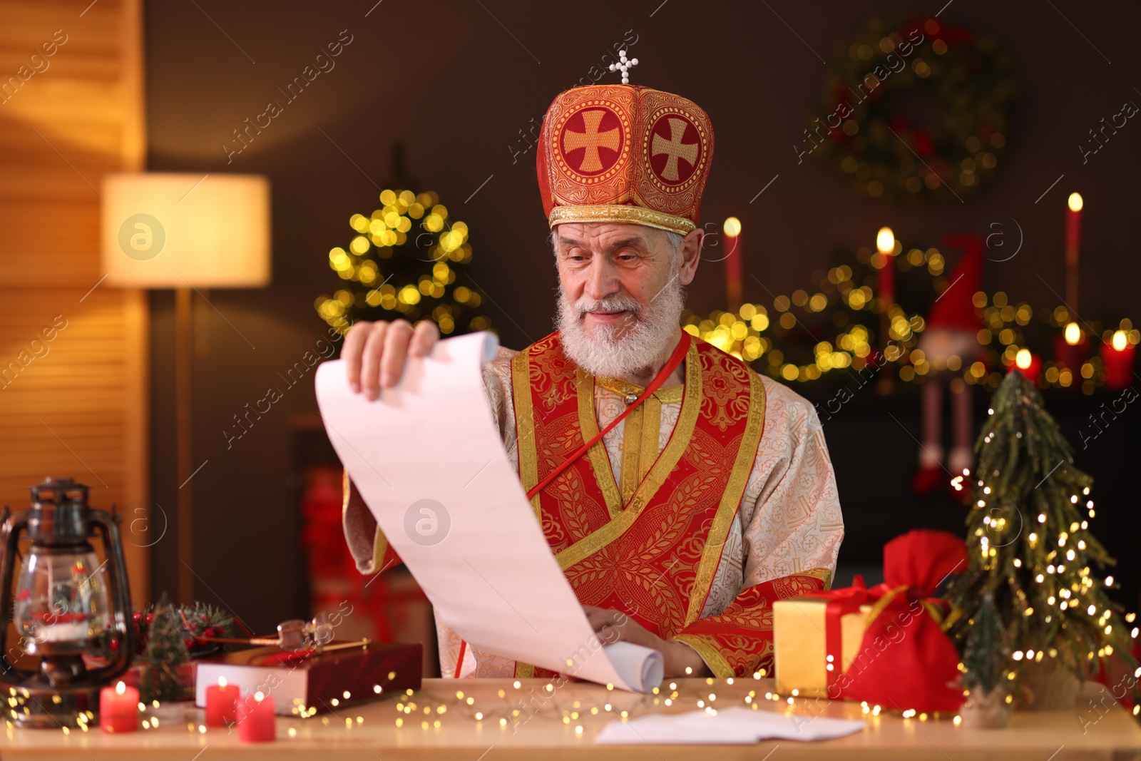 Photo of Saint Nicholas with gift list at desk in room decorated for Christmas