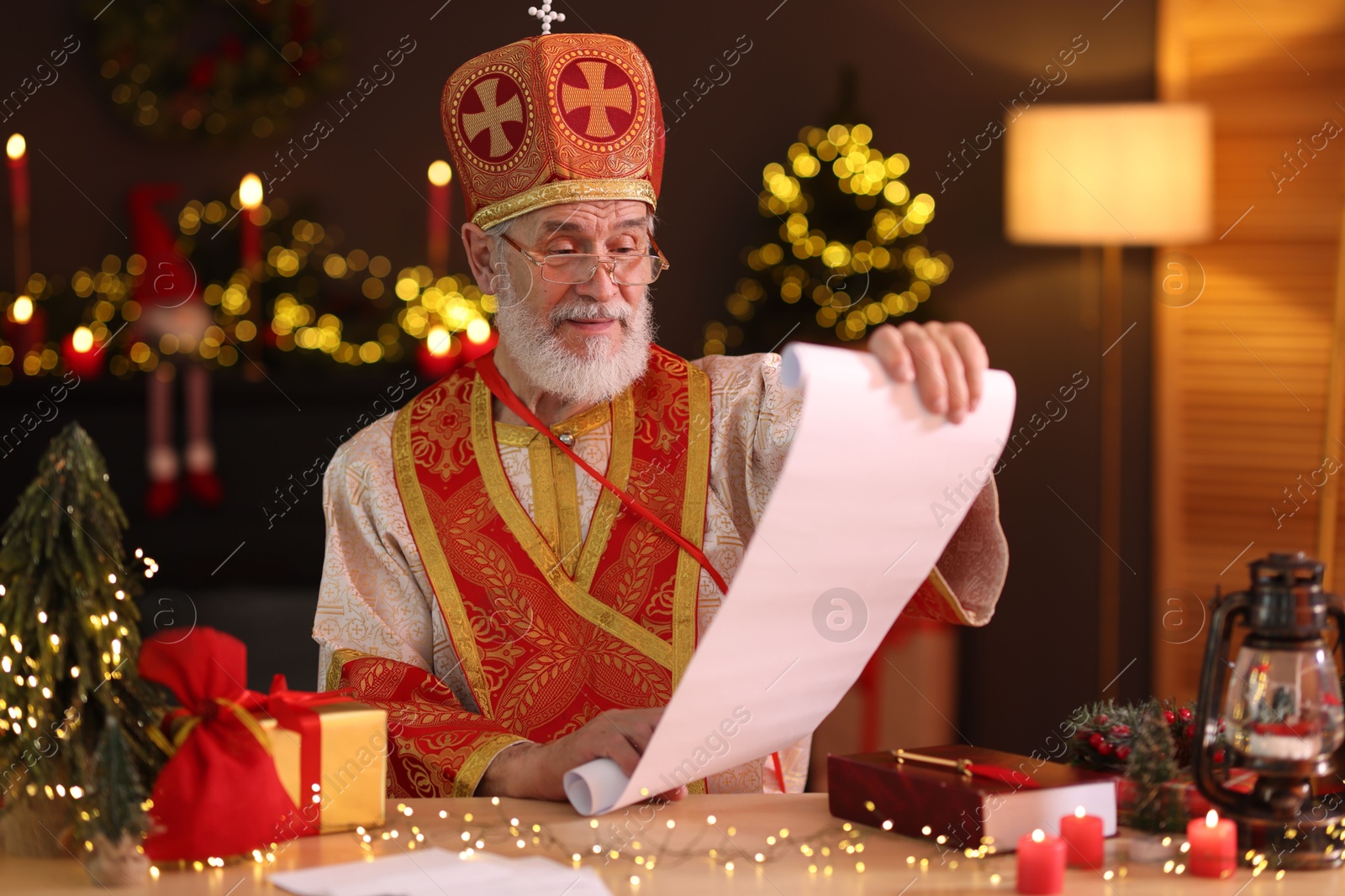 Photo of Saint Nicholas with gift list at desk in room decorated for Christmas