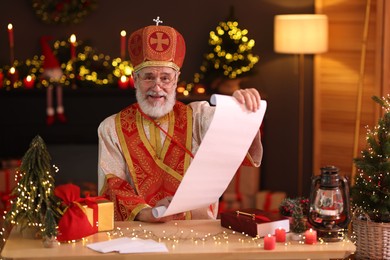 Photo of Saint Nicholas with gift list at desk in room decorated for Christmas