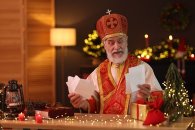 Saint Nicholas with letters at desk in room decorated for Christmas