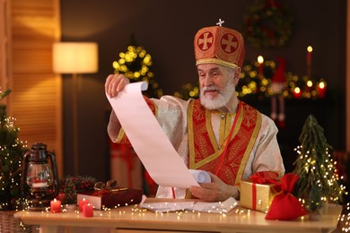 Photo of Saint Nicholas with gift list at desk in room decorated for Christmas