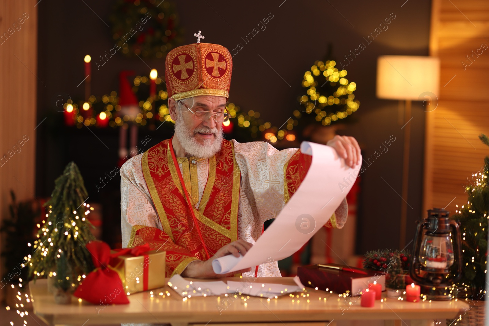 Photo of Saint Nicholas with gift list at desk in room decorated for Christmas