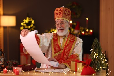 Saint Nicholas with gift list at desk in room decorated for Christmas