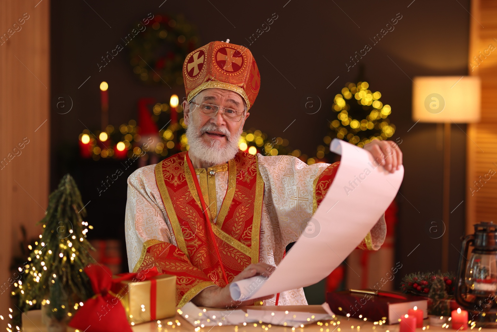 Photo of Saint Nicholas with gift list at desk in room decorated for Christmas