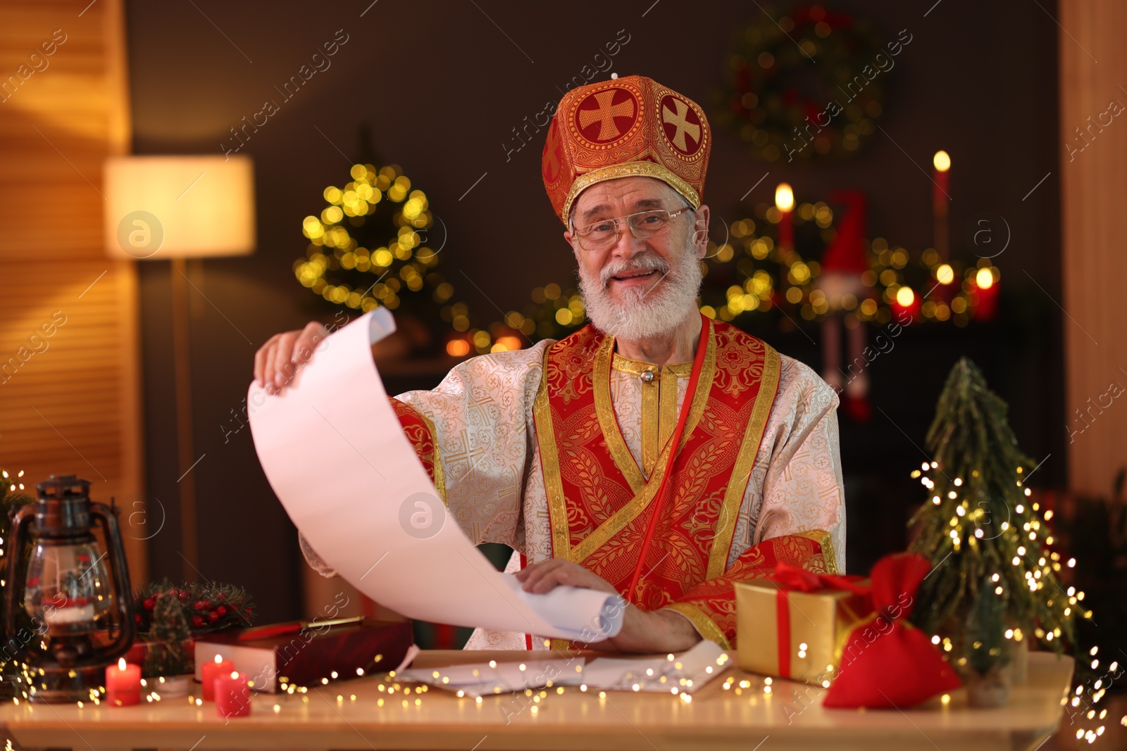 Photo of Saint Nicholas with gift list at desk in room decorated for Christmas