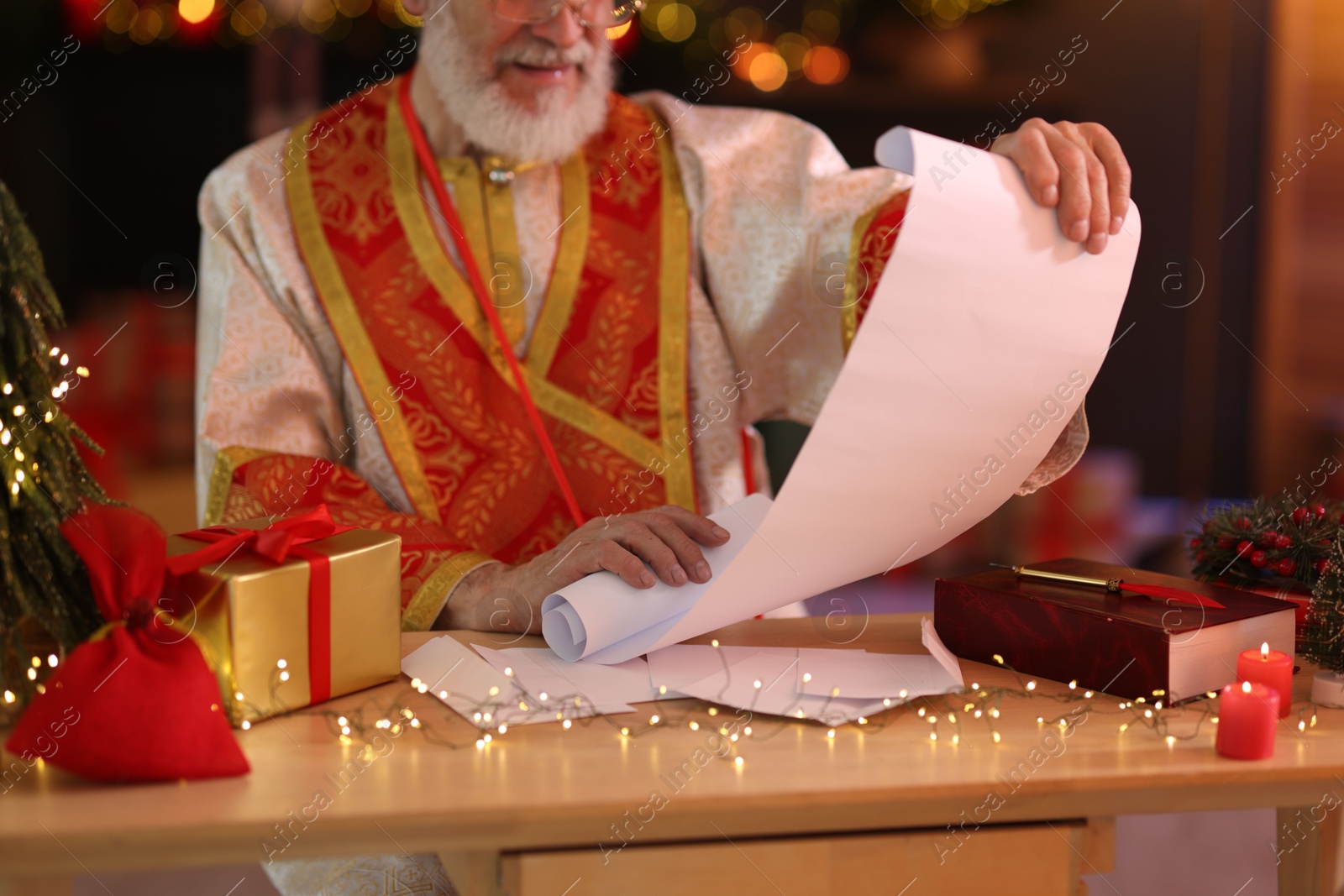 Photo of Saint Nicholas with gift list at desk in room decorated for Christmas, closeup