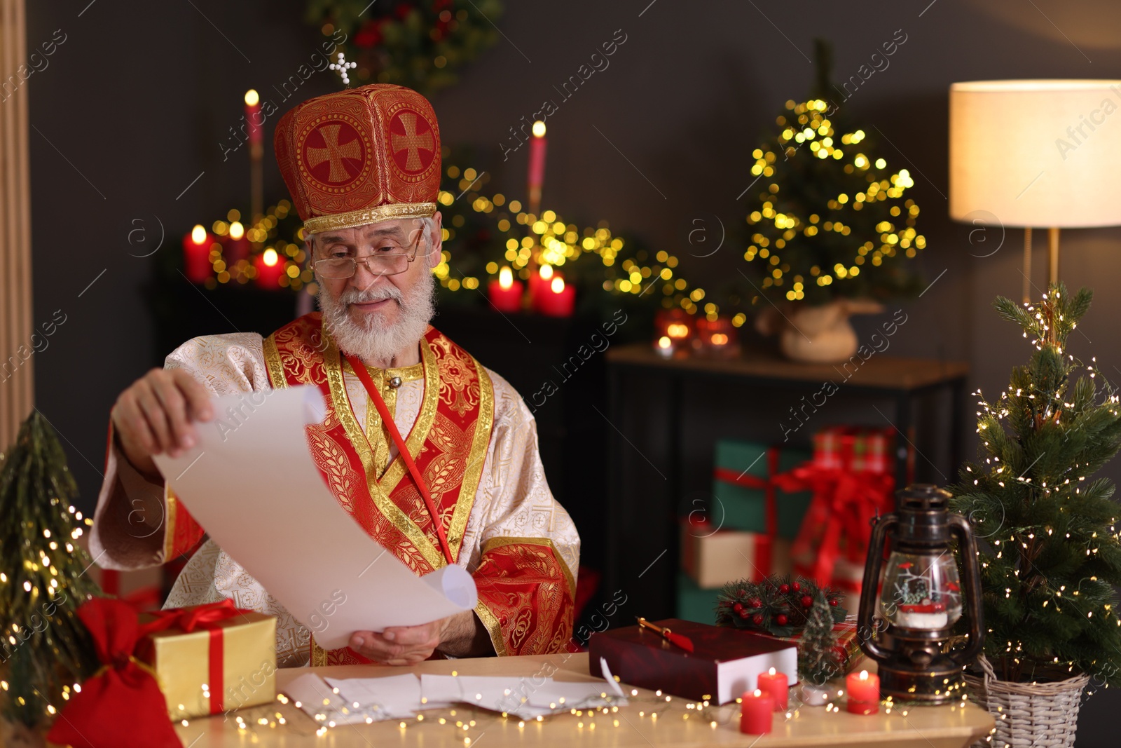 Photo of Saint Nicholas with gift list at desk in room decorated for Christmas