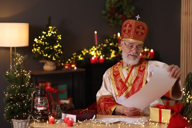 Photo of Saint Nicholas with gift list at desk in room decorated for Christmas