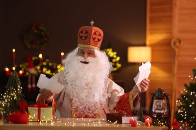 Saint Nicholas with letters at desk in room decorated for Christmas