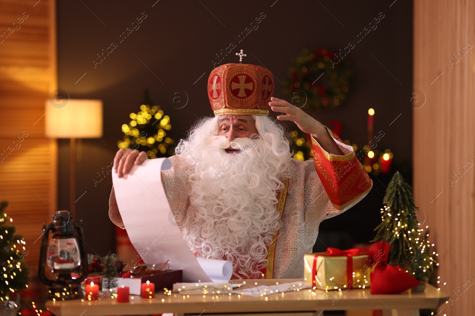 Photo of Saint Nicholas with gift list at desk in room decorated for Christmas