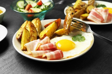 Photo of Tasty brunch. Fried egg, potato, bacon and fork on dark textured table, closeup