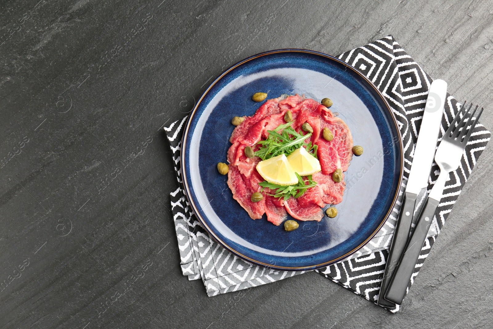 Photo of Fresh beef carpaccio with arugula, lemon and capers on dark textured table, top view. Space for text