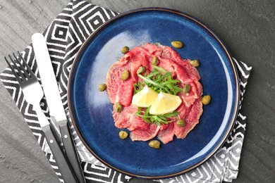 Photo of Fresh beef carpaccio with arugula, lemon and capers on dark textured table, top view