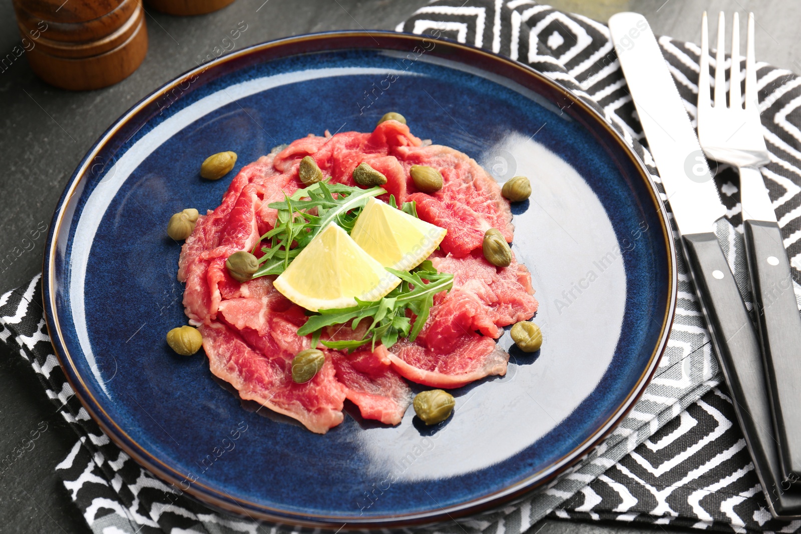 Photo of Fresh beef carpaccio with arugula, lemon and capers on dark textured table, closeup