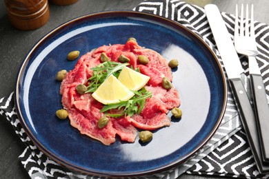 Photo of Fresh beef carpaccio with arugula, lemon and capers on dark textured table, closeup