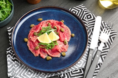 Photo of Fresh beef carpaccio with arugula, lemon and capers on dark textured table, above view