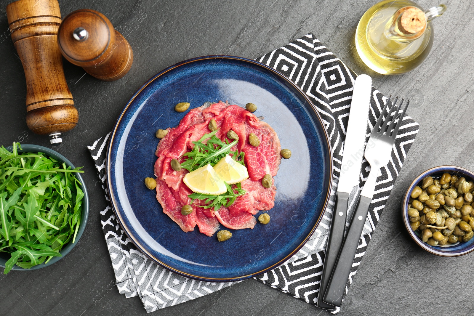 Photo of Fresh beef carpaccio with arugula, lemon and capers served on dark textured table, flat lay