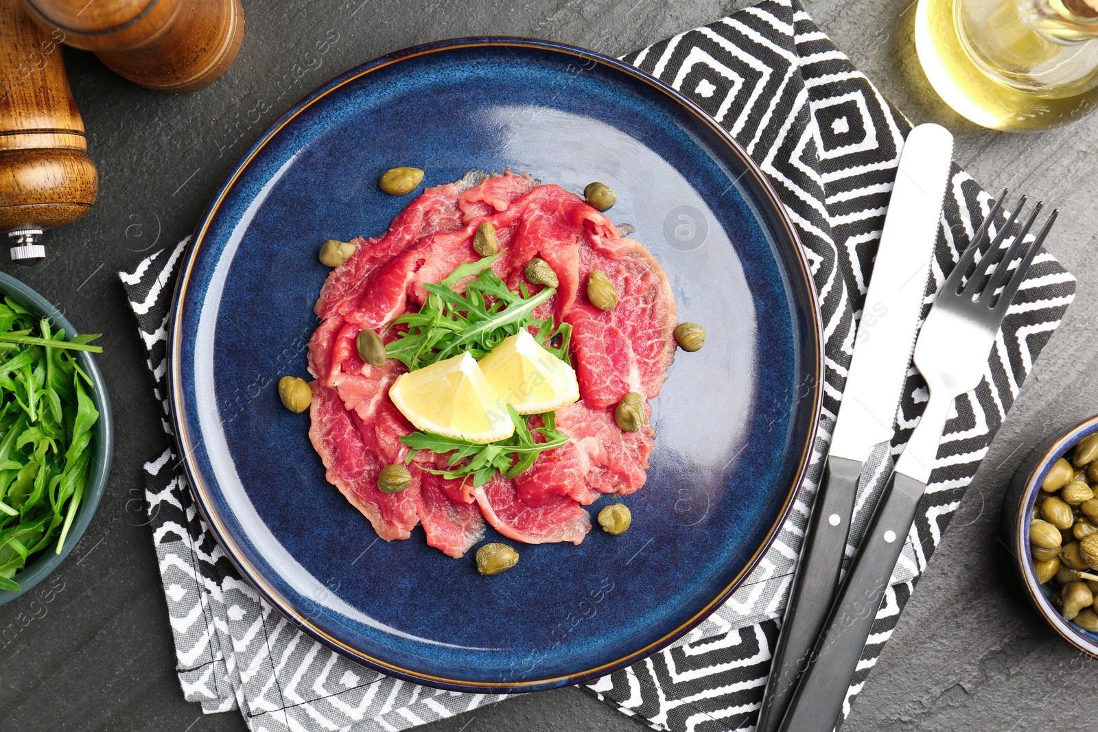 Photo of Fresh beef carpaccio with arugula, lemon and capers served on dark textured table, flat lay