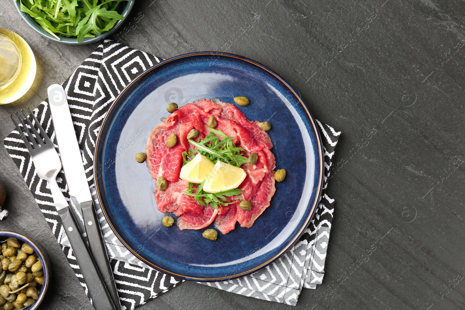 Photo of Fresh beef carpaccio with arugula, lemon and capers served on dark textured table, flat lay. Space for text