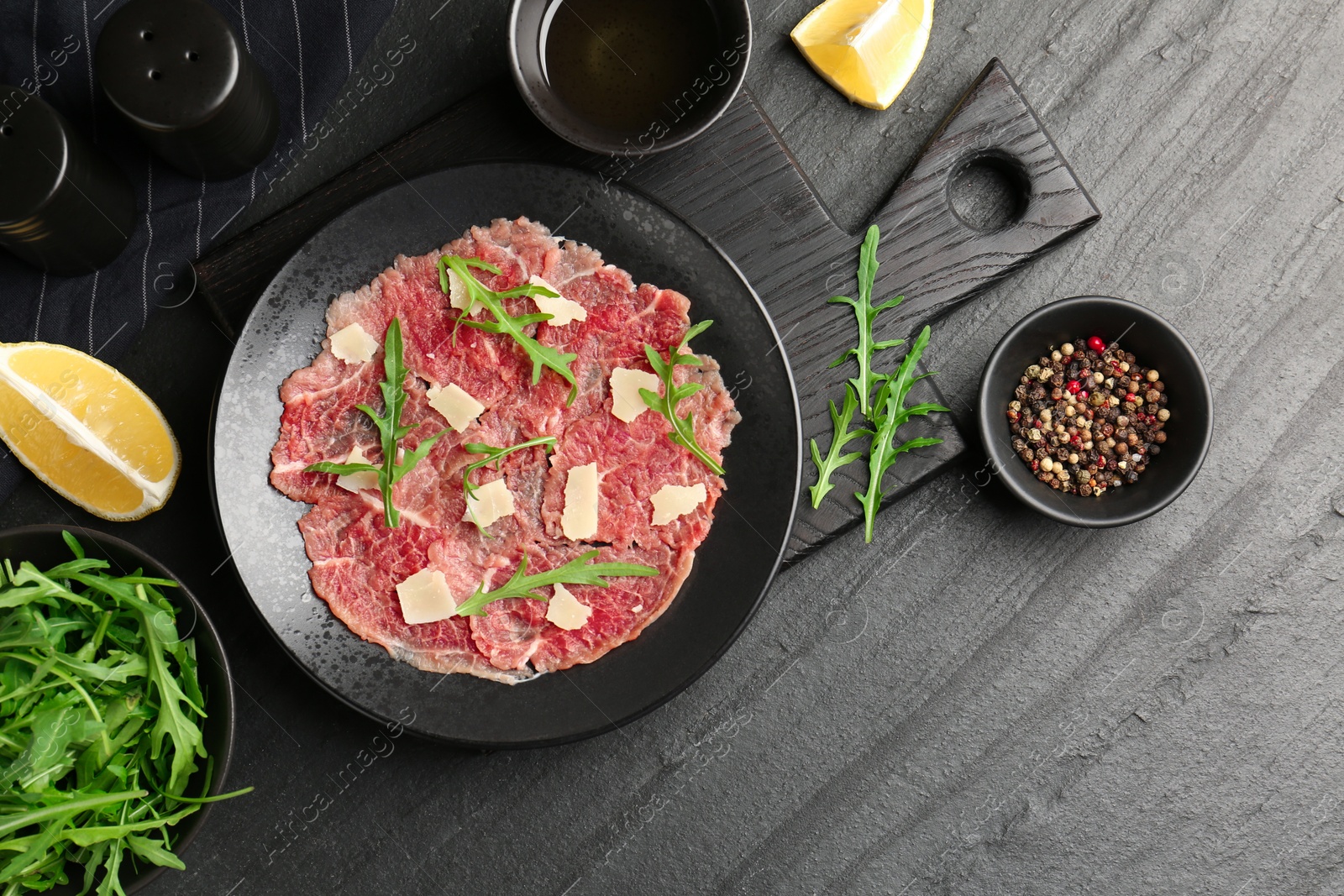 Photo of Fresh beef carpaccio with arugula and cheese on dark textured table, flat lay. Space for text