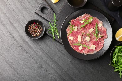 Photo of Fresh beef carpaccio with arugula and cheese on dark textured table, flat lay. Space for text