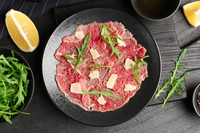 Photo of Fresh beef carpaccio with arugula and cheese on dark textured table, flat lay